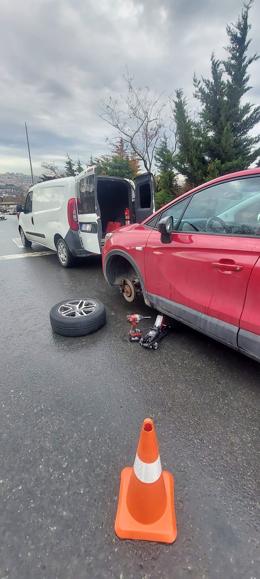 Yol kenarında kırmızı aracın lastiğinin kriko ve somun sökücüyle değiştirilmesi, yanında trafik konisi